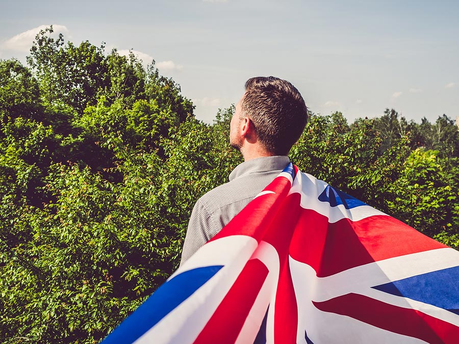 Attractive man waving a British Flag against a background of trees and blue sky. View from the back, close-up. National holiday concept